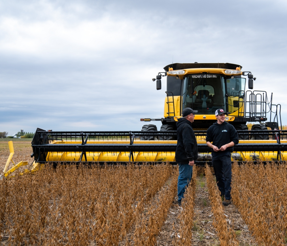 Two farmers discuss in front of a yellow combine harvester during the harvest of soybeans.